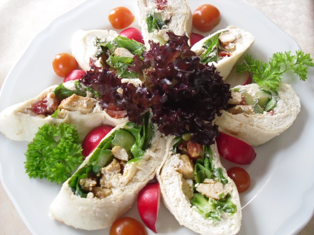 Dekoracyjnie ułożone roladki z tortilli z kurczakiem, warzywami i serem, otoczone rzodkiewkami i pomidorkami cherry na białym talerzu.