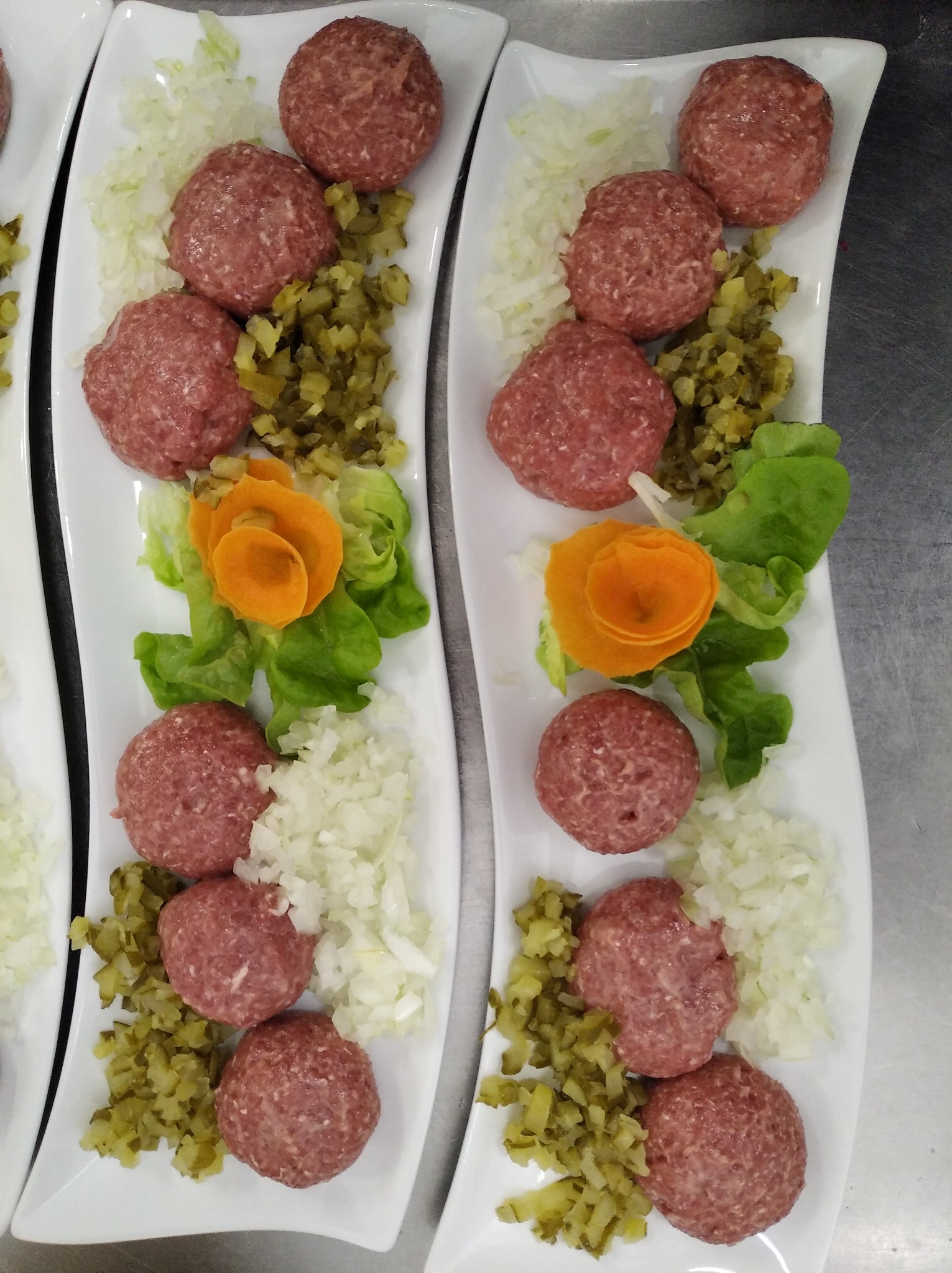 Białe, prostokątne talerze z trzema surowymi kulkami mięsnymi, posiekaną cebulą, korniszonami i dekoracją z marchewki w kształcie różyczki, przygotowywane do dalszej obróbki.