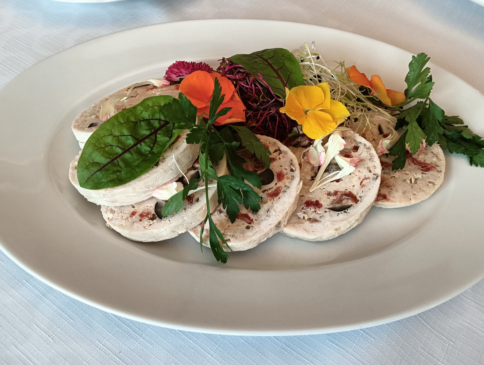 Dekoracyjnie ułożone plastry pasztetu z dodatkiem ziół i jadalnych kwiatów na białym talerzu, na tle białego obrusu.