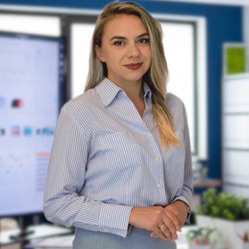 Portret kobiety w koszuli w paski, z dłońmi splecionymi przed sobą, w tle biuro z monitorem wyświetlającym wykresy.