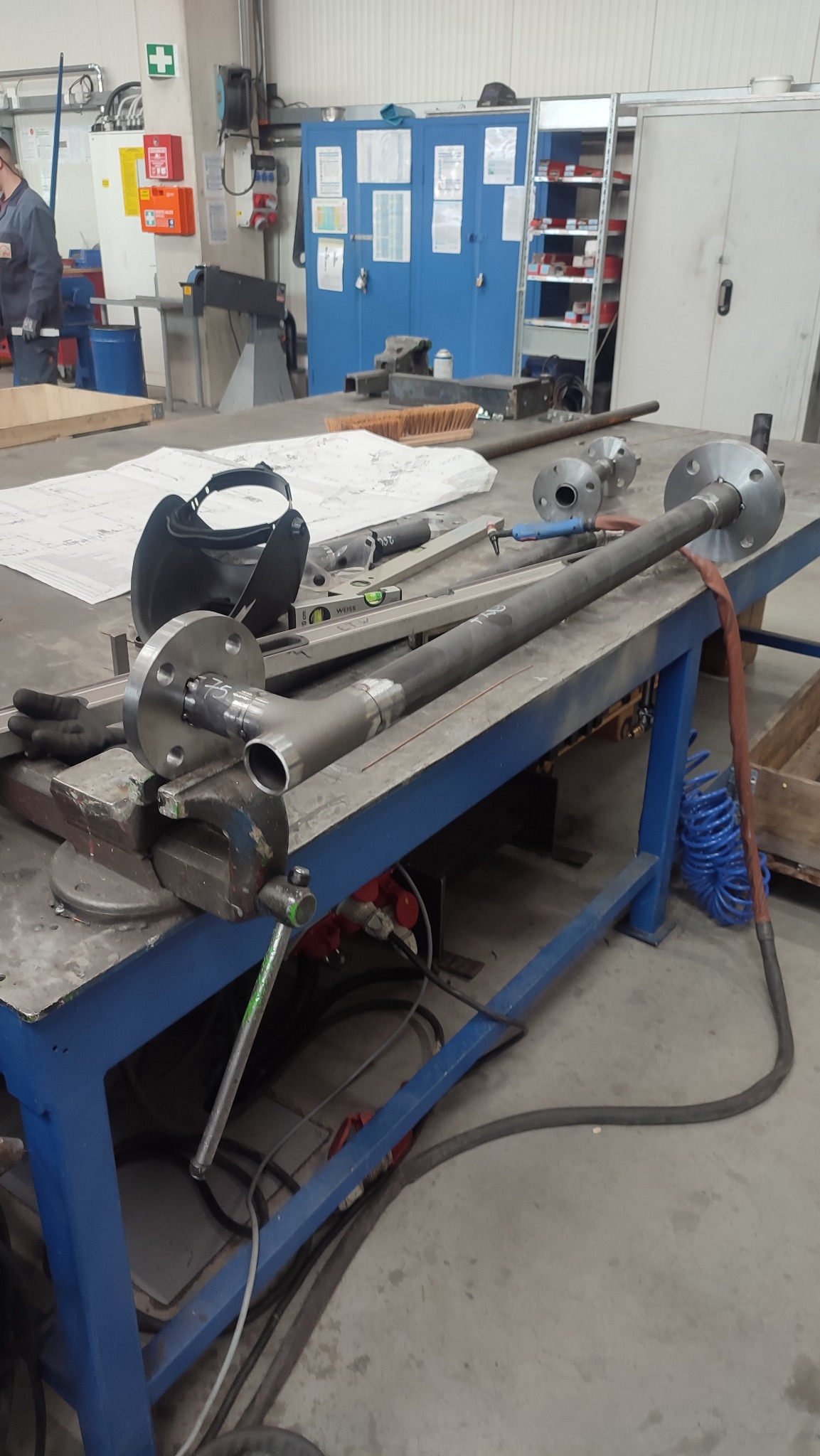 Element rurociągu z przyspawanymi kołnierzami, mocowany w imadle na stole warsztatowym; obok leży maska spawalnicza i rysunek techniczny.