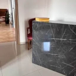 Recepcja wykończona czarnym marmurem z białymi żyłkami, na blacie leży duża, złota książka, w tle czerwone krzesło i widok na salon.