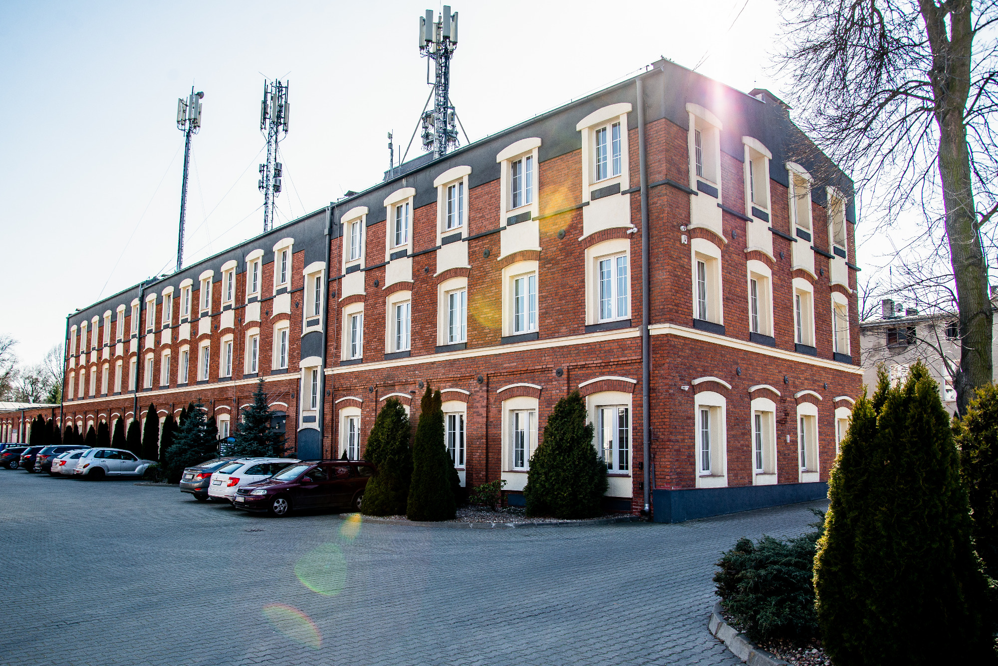 Ceglany budynek biurowy z rzędami okien, antenami na dachu i parkingiem z samochodami przed nim.