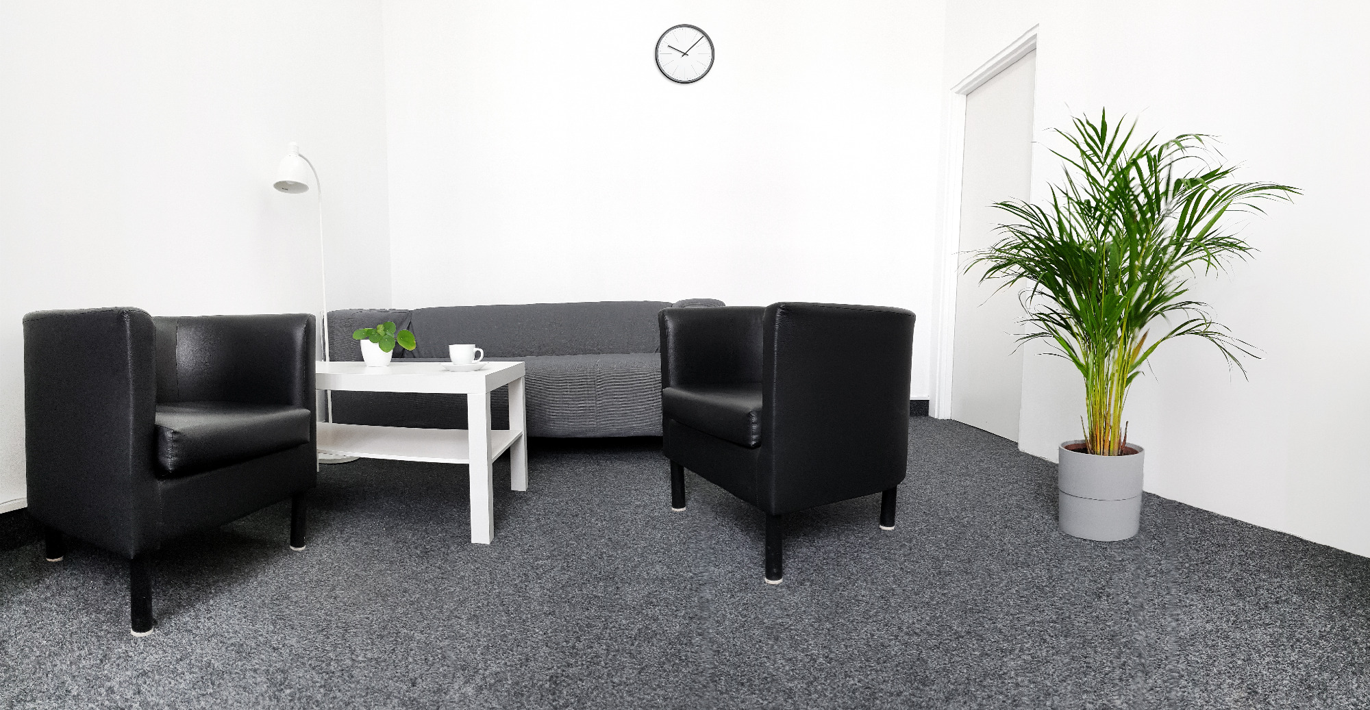 Jasne, minimalistyczne wnętrze gabinetu z dwoma czarnymi fotelami, szarą sofą, białym stolikiem z rośliną i filiżanką, zegarem na ścianie i dużą rośliną doniczkową.