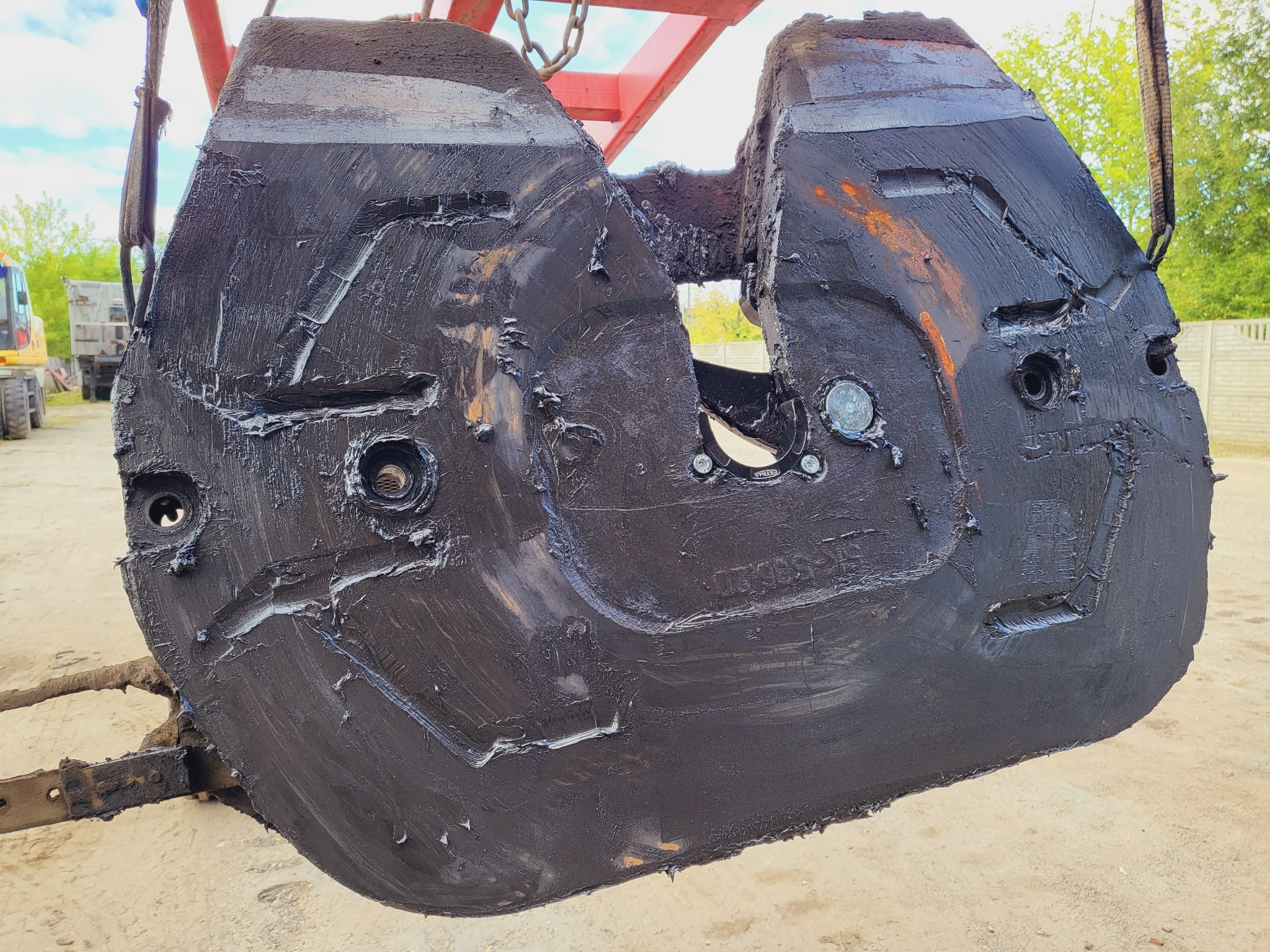 Ciężki, brudny element maszyny budowlanej, prawdopodobnie łyżka koparki, zawieszony na łańcuchach podnośnika, z widocznymi śladami smaru i zużycia.