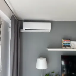 Biała jednostka klimatyzacyjna zamontowana na szarej ścianie w salonie, widoczna roleta okienna, zasłona, lampa podłogowa i półka z książkami