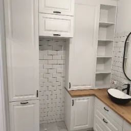 Biała zabudowa łazienkowa z drewnianym blatem, umywalką i półkami, wykończona białymi płytkami typu metro. Minimalistyczny design z czarnymi uchwytami.