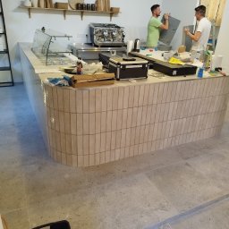 One Service Jakub Małecki - Montaż lady barowej w kawiarni: beżowe płytki, ekspres do kawy, półki z dekoracjami. Dwóch monterów pracuje nad wykończeniem, widoczne narzędzia i akcesoria.