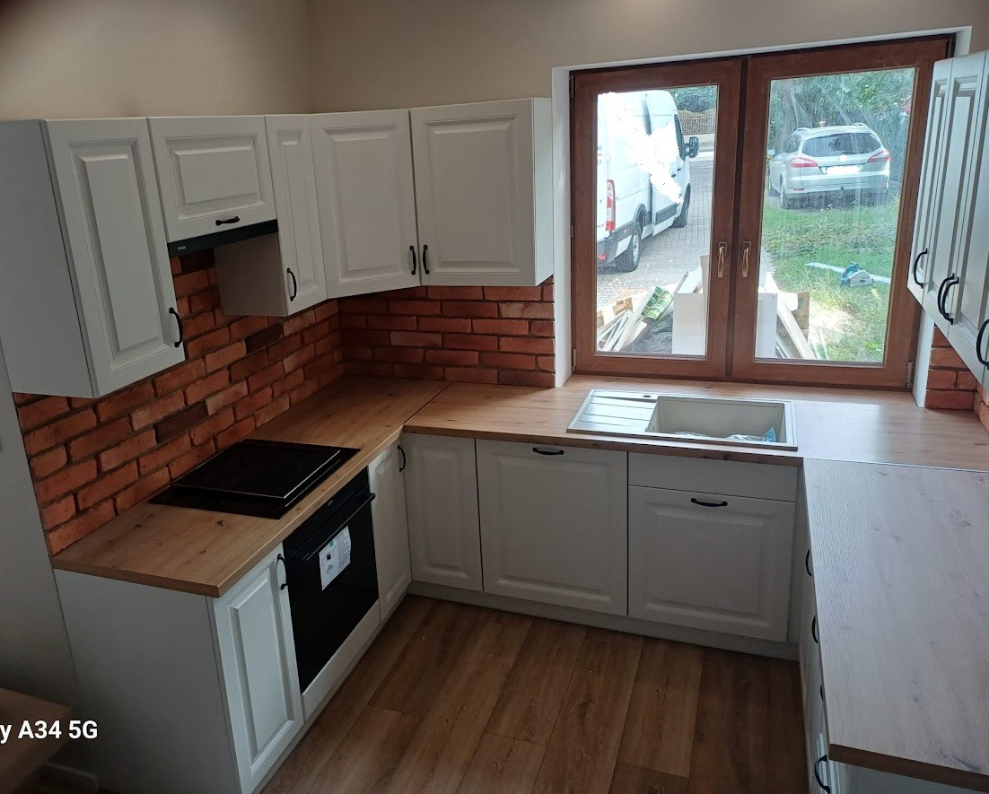 Biała kuchnia z drewnianym blatem i ceglaną ścianą. Widok na okno z zewnątrz. Nowoczesny design z elementami rustykalnymi. Ustawienie w kształcie litery 'U'.