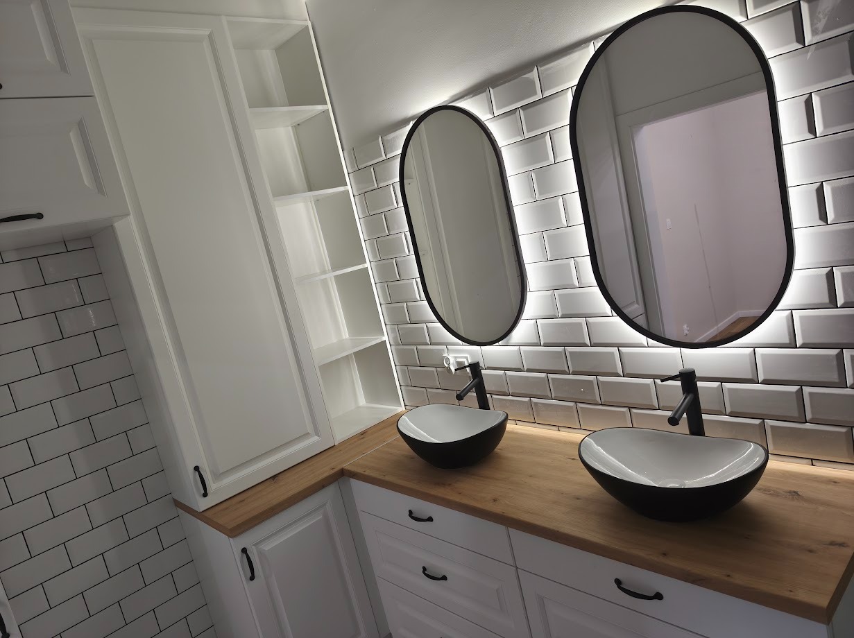 Biała łazienka z dwoma umywalkami w czarnych misach i podświetlanymi lustrami owalnymi, ułożonymi na tle białych płytek typu metro, z drewnianym blatem i szafkami.