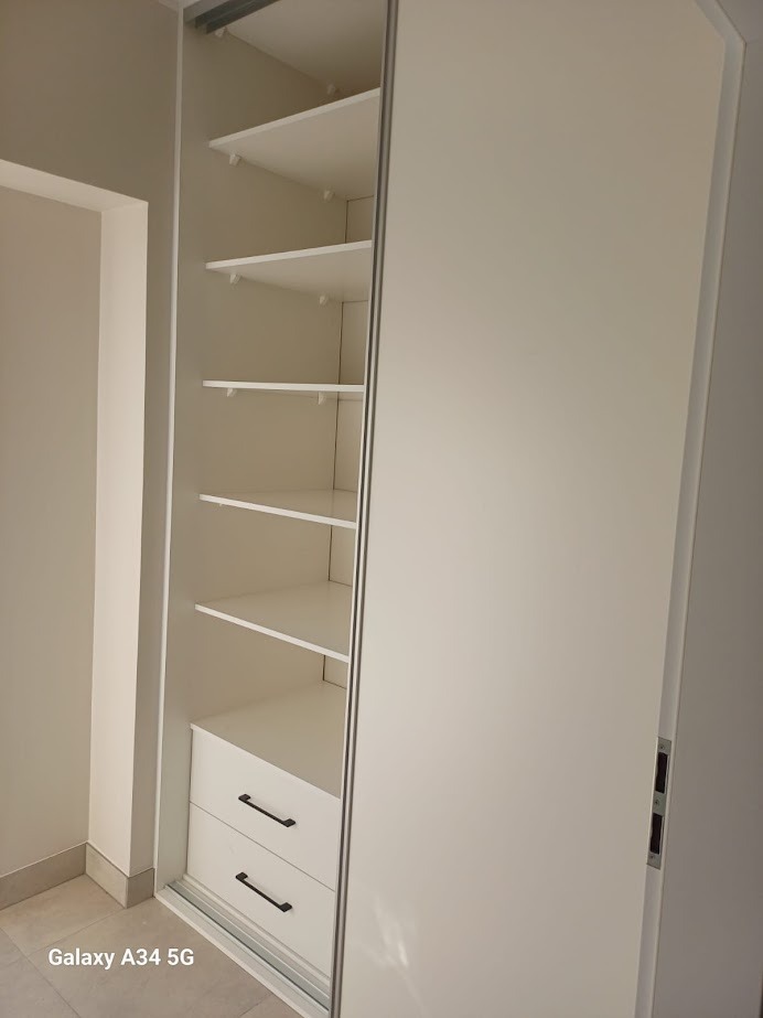 Biała szafa wnękowa z przesuwnymi drzwiami, częściowo otwarta, ukazująca półki i dwie szuflady z czarnymi uchwytami. Minimalistyczny design, jasne wnętrze.