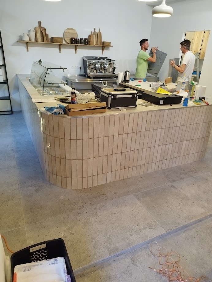 Montaż lady barowej w kawiarni: beżowe płytki, ekspres do kawy, półki z dekoracjami. Dwóch monterów pracuje nad wykończeniem, widoczne narzędzia i akcesoria.