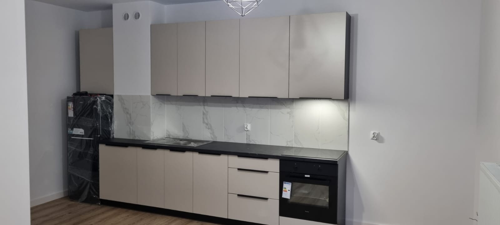 Jasna kuchnia z beżowymi szafkami i czarnym blatem. Nowa lodówka w folii ochronnej. Marmurowe płytki nad blatem. Minimalistyczny design, nowoczesne oświetlenie.
