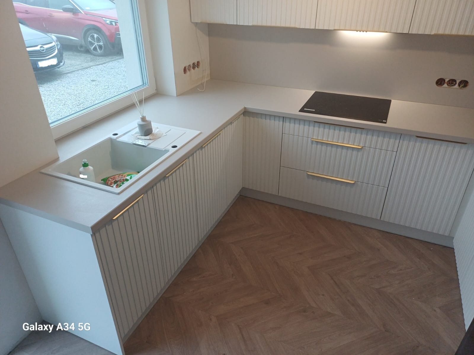 Jasna kuchnia na wymiar z frezowanymi frontami, złotymi uchwytami i drewnianą podłogą w jodełkę. Widok na zlew, płytę indukcyjną i okno z widokiem na samochody.