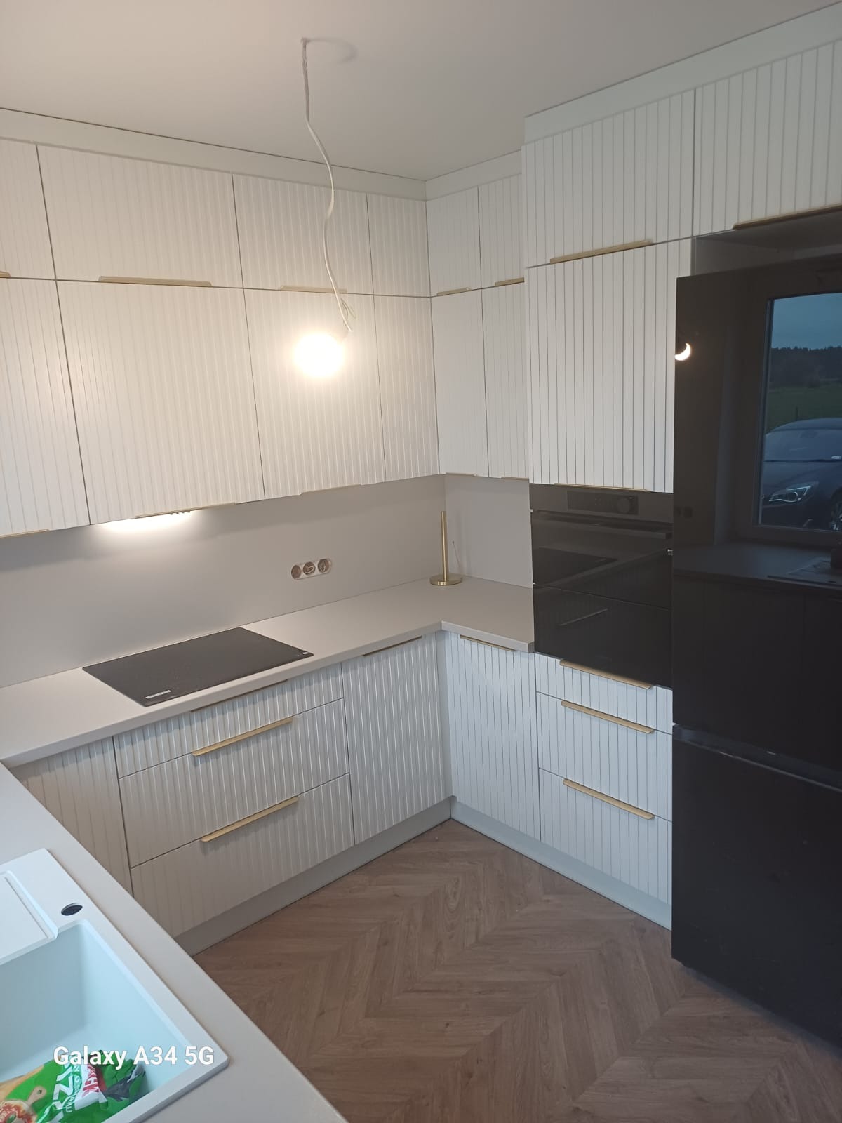 Biała kuchnia na wymiar z frezowanymi frontami i złotymi uchwytami, parkiet w jodełkę. Widoczny fragment zlewu i czarny sprzęt AGD.