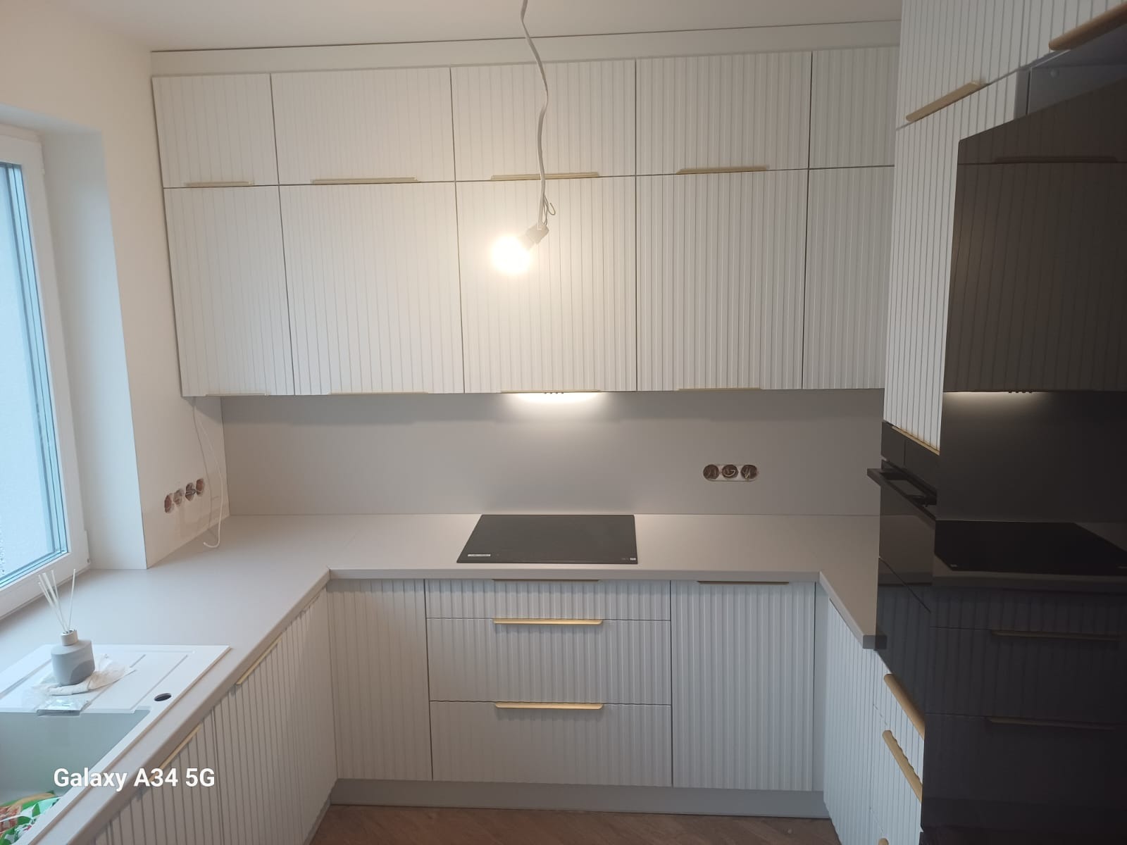 Jasna kuchnia na wymiar z frezowanymi frontami i złotymi uchwytami. Widoczna płyta indukcyjna i okno z dyfuzorem zapachowym. Minimalistyczny design.