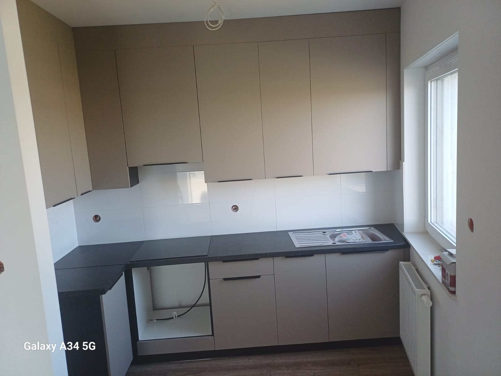 Jasna kuchnia z szarym blatem i białymi płytkami, widok na szafki wiszące i dolne, zlew oraz fragment okna z grzejnikiem. Część szafek bez frontów.