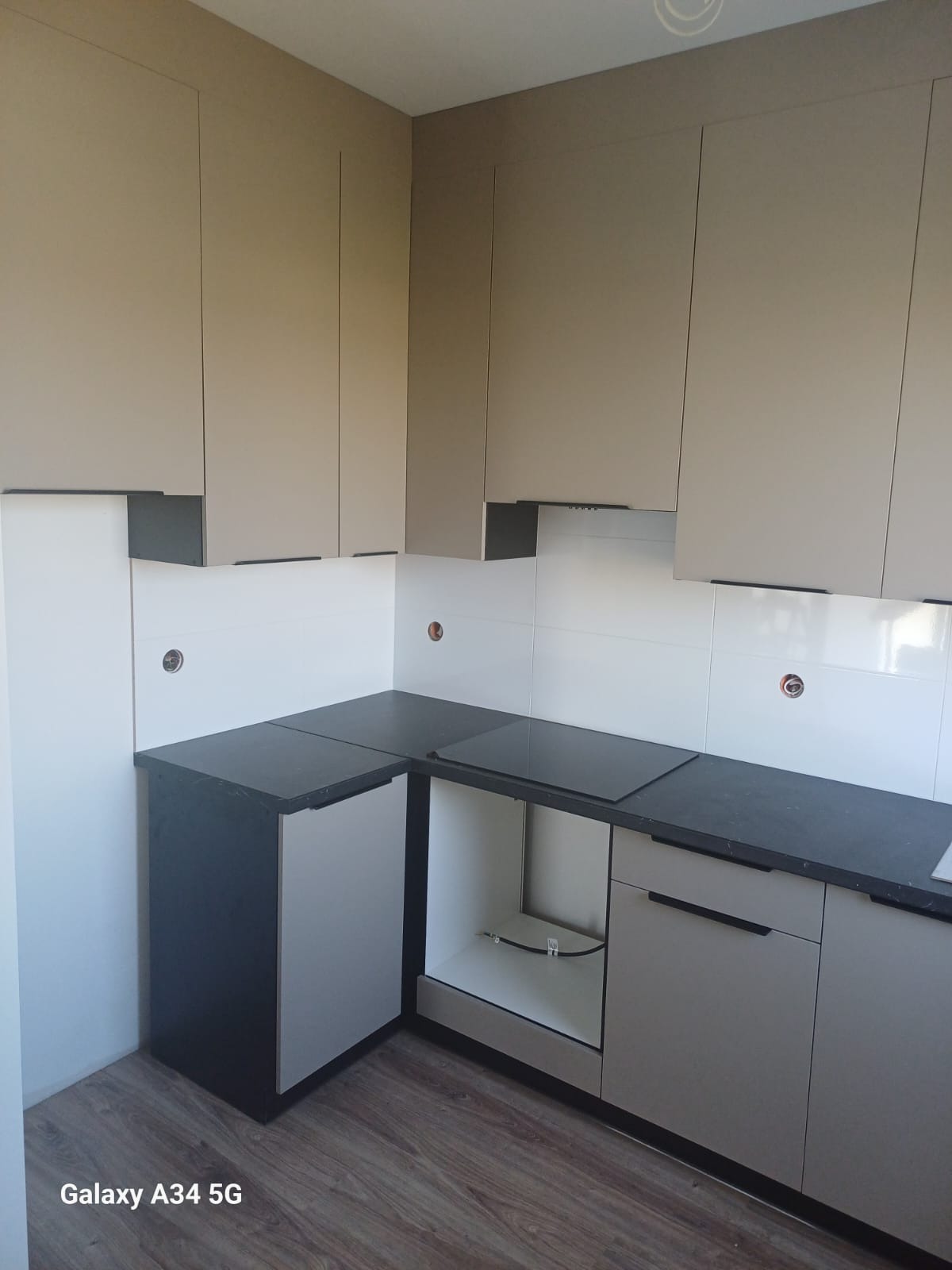 Nowoczesna kuchnia z beżowymi szafkami górnymi i ciemnymi blatami. Widoczne białe płytki na ścianie i drewniana podłoga. Część szafek dolnych bez frontów.