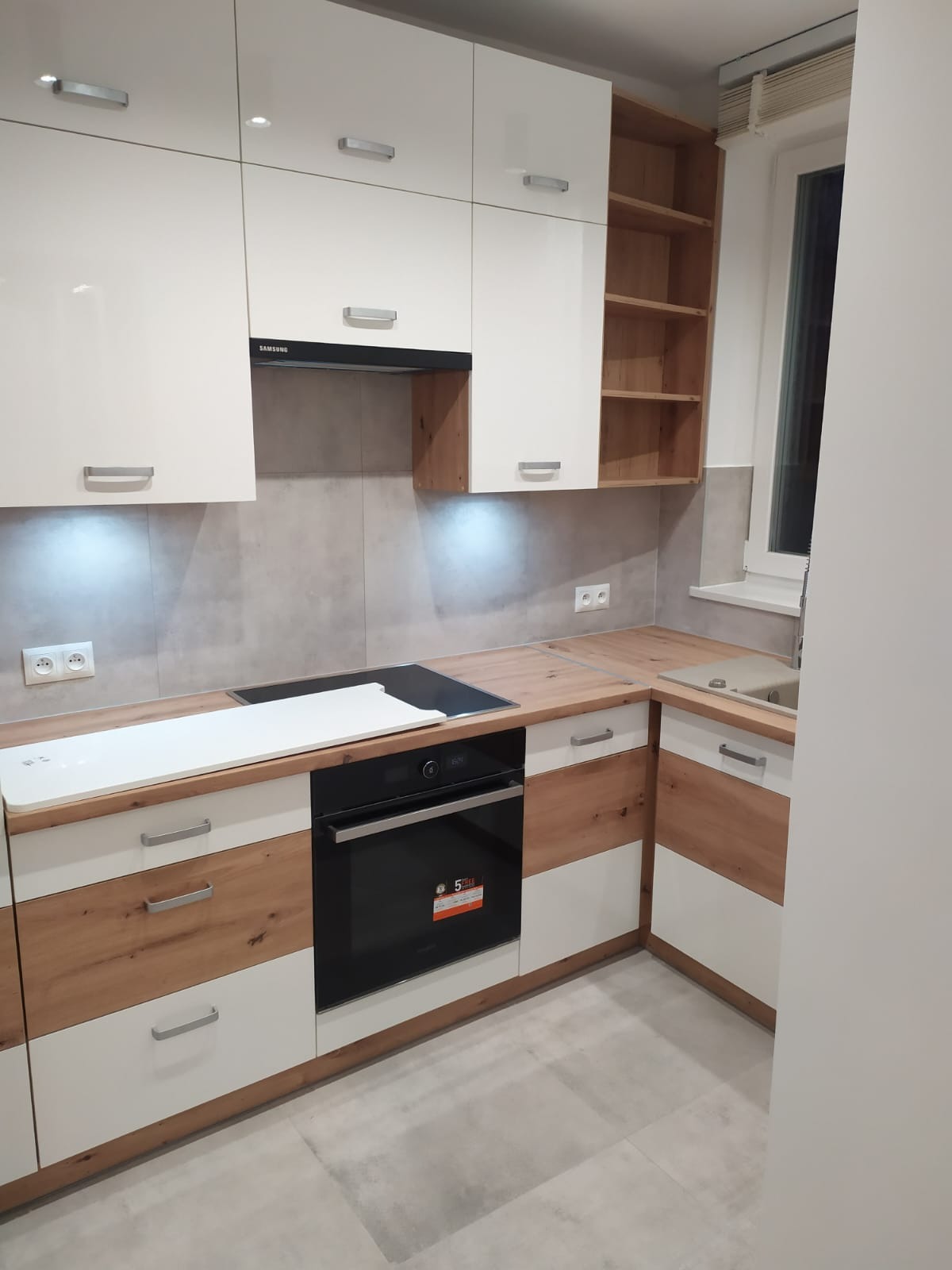 Nowoczesna kuchnia z białymi szafkami górnymi i drewnopodobnymi dolnymi, czarny piekarnik, szara ściana i blat. Minimalistyczny design, jasne oświetlenie.