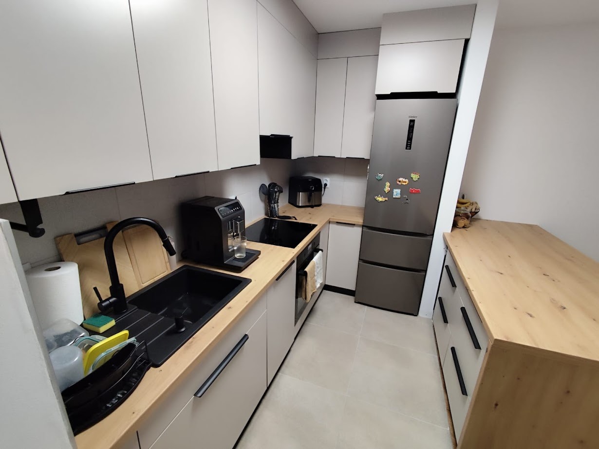 Jasna kuchnia na wymiar z drewnianym blatem, czarnym zlewem i srebrną lodówką. Minimalistyczny design, zabudowa pod sufit, nowoczesne wykończenie.