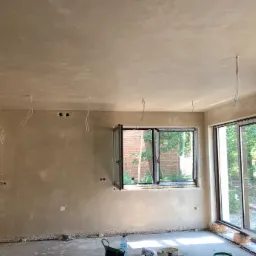 Świeżo położone tynki maszynowe na ścianach i suficie w jasnym pokoju z otwartym oknem i dużym oknem przesuwnym, widoczne wystające kable elektryczne i wiadro na podłodze.