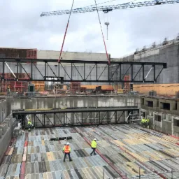 Montaż stalowej konstrukcji nośnej na placu budowy, widoczne dźwigi, robotnicy w odblaskowych kamizelkach i kaskach, szalunki oraz betonowe ściany w surowym stanie.