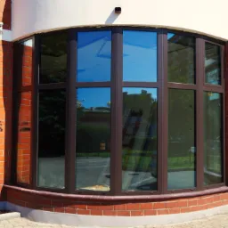 Zaokrąglone okno z brązowymi ramami w budynku z czerwonej cegły, widoczne odbicia otoczenia w szybach.