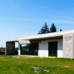 Surowy stan domu jednorodzinnego z zamontowanymi oknami i drzwiami, widoczne samochody i spacerujące osoby na tle błękitnego nieba.