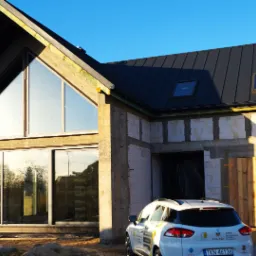 Surowy stan domu jednorodzinnego z dużymi przeszkleniami, widoczna brama garażowa z surowego drewna, biały samochód firmowy na podjeździe, dach pokryty blachą na rąbek stojący.