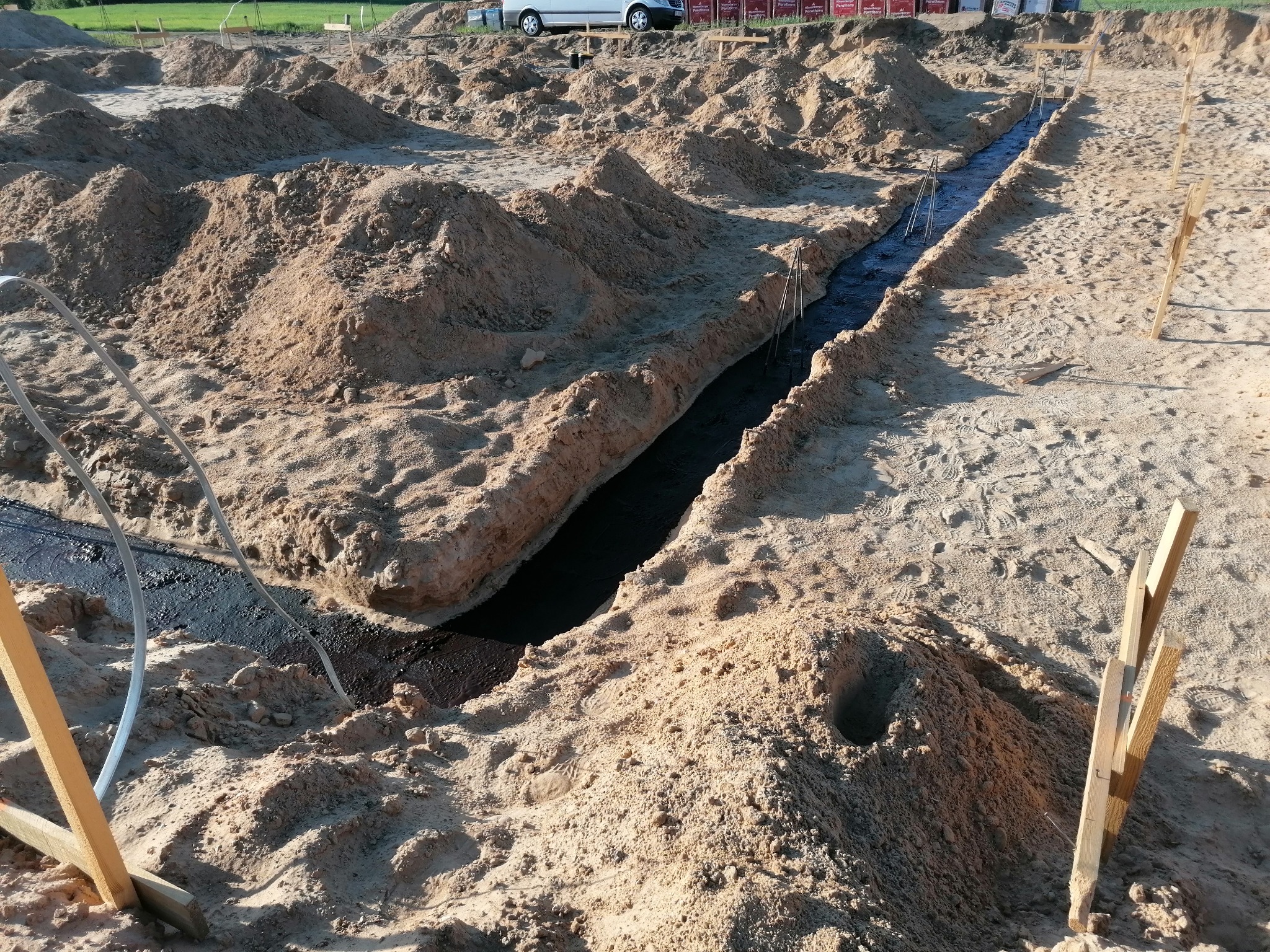 Wykop fundamentowy wypełniony czarną folią izolacyjną, otoczony piaskiem i drewnianymi palikami, widoczne ślady stóp na piasku, w tle zaparkowany biały samochód dostawczy