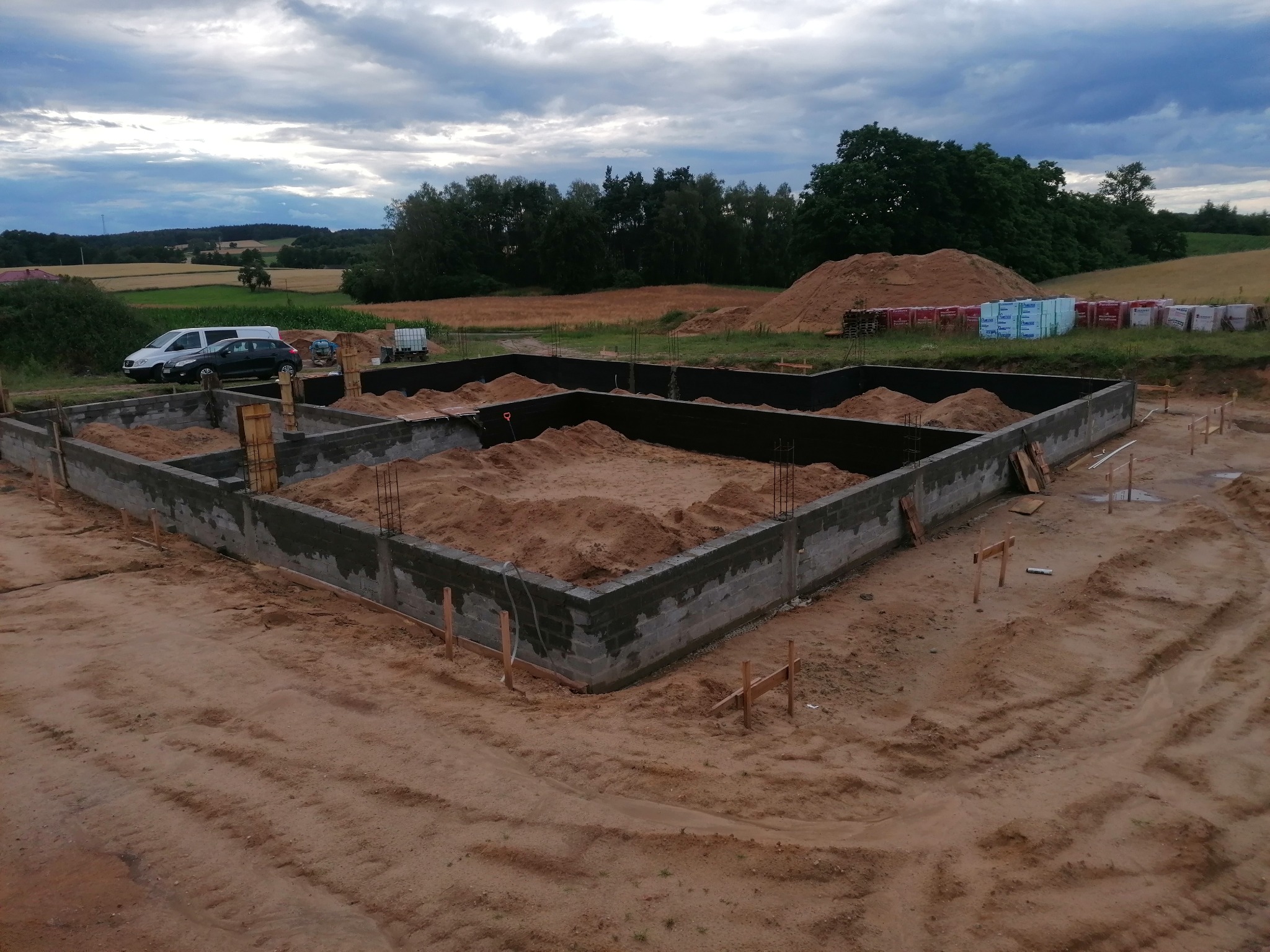 Fundament budynku z izolacją przeciwwilgociową, zasypany piaskiem, widoczne zbrojenia, teren budowy z zaparkowanymi samochodami i składowanym materiałem budowlanym w tle.