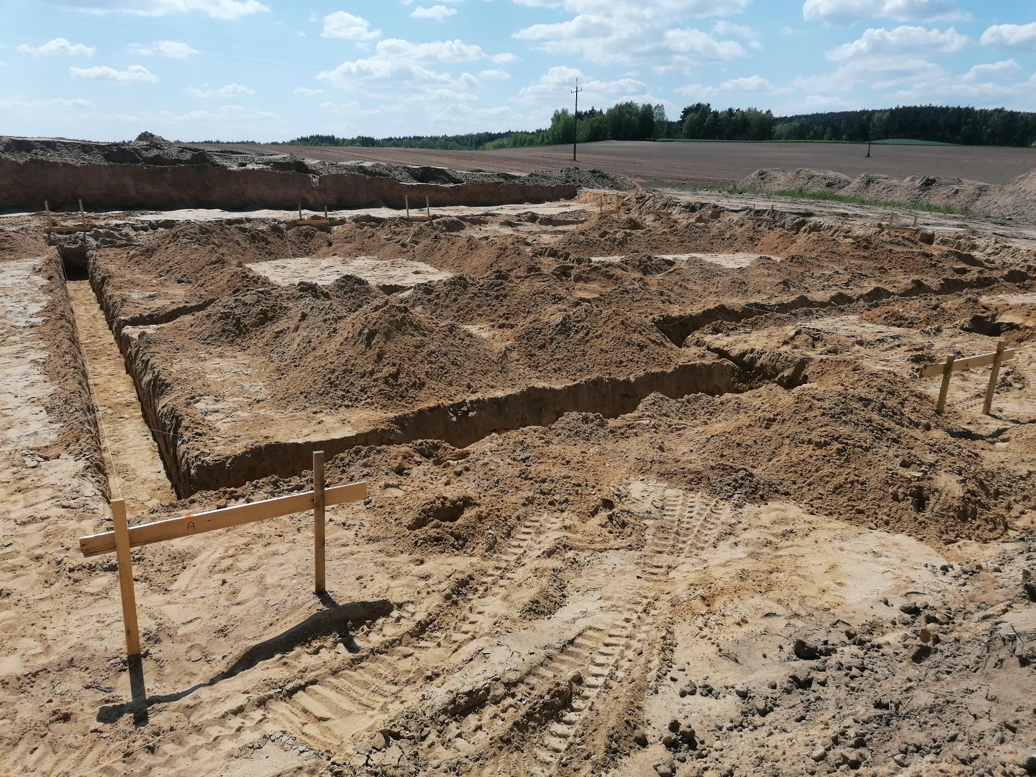 Wykonane wykopy pod fundamenty budynku z drewnianymi stemplami i wytyczonym obrysem, widoczne ślady opon na piaszczystym podłożu, w tle pole uprawne i las.