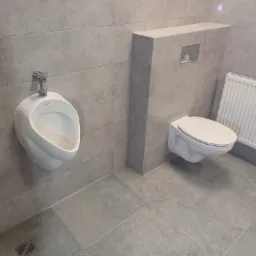 Szary, wykafelkowany pokój z zamontowanym pisuarami i toaletą podwieszaną z grzejnikiem.