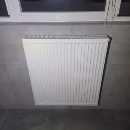 Biały, nowy grzejnik panelowy zamontowany na szarej, kafelkowej ścianie, widok z dołu, z widocznym fragmentem okna i ciemnym otoczeniem za nim.