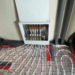 Rozdzielacz ogrzewania podłogowego z podłączonymi rurami na folii izolacyjnej z nadrukiem, widoczne szare rury instalacyjne przy ścianie.