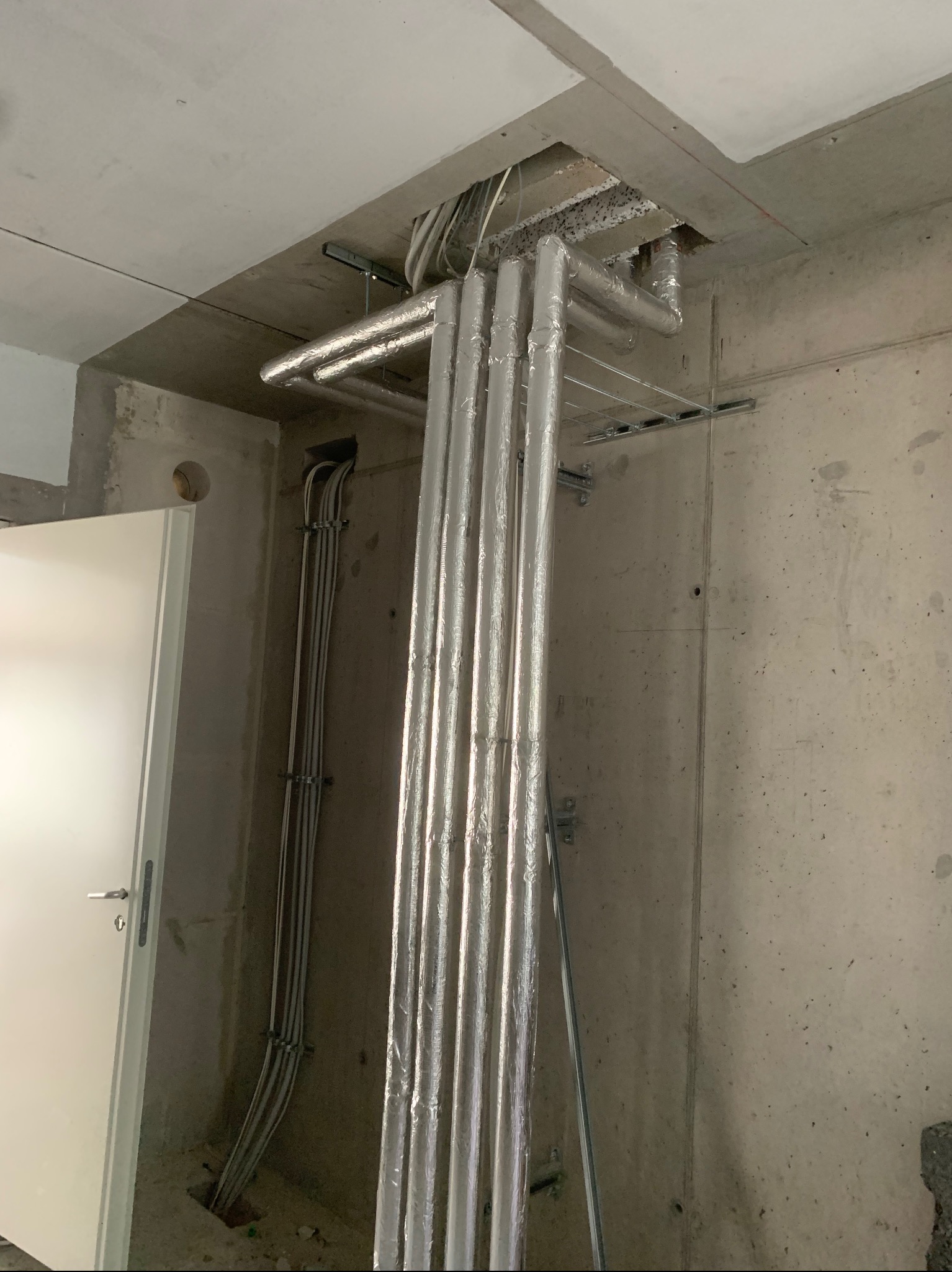 Rury grzewcze w srebrnej izolacji, prowadzone pionowo w surowym wnętrzu w trakcie instalacji, widoczne mocowania do sufitu i ściany, oraz kable elektryczne w peszlach.