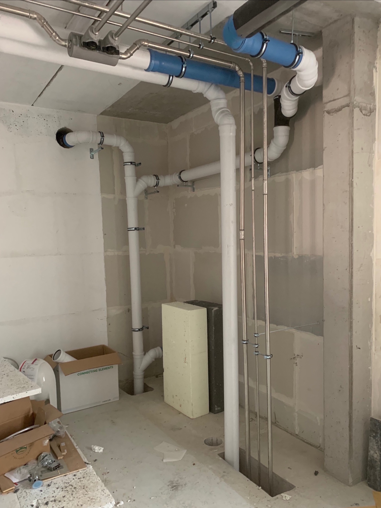 Surowe wnętrze w trakcie instalacji: widoczne rury kanalizacyjne i grzewcze w różnych kolorach (biały, niebieski, czarny) oraz metalowe rurki, prowadzone wzdłuż ściany. W tle surowe ściany...