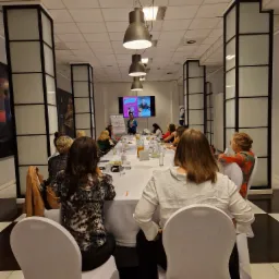 Sala konferencyjna z uczestnikami szkolenia siedzącymi przy długim stole, prelegentka prezentuje materiał na ekranie, widoczne notatki na flipcharcie.