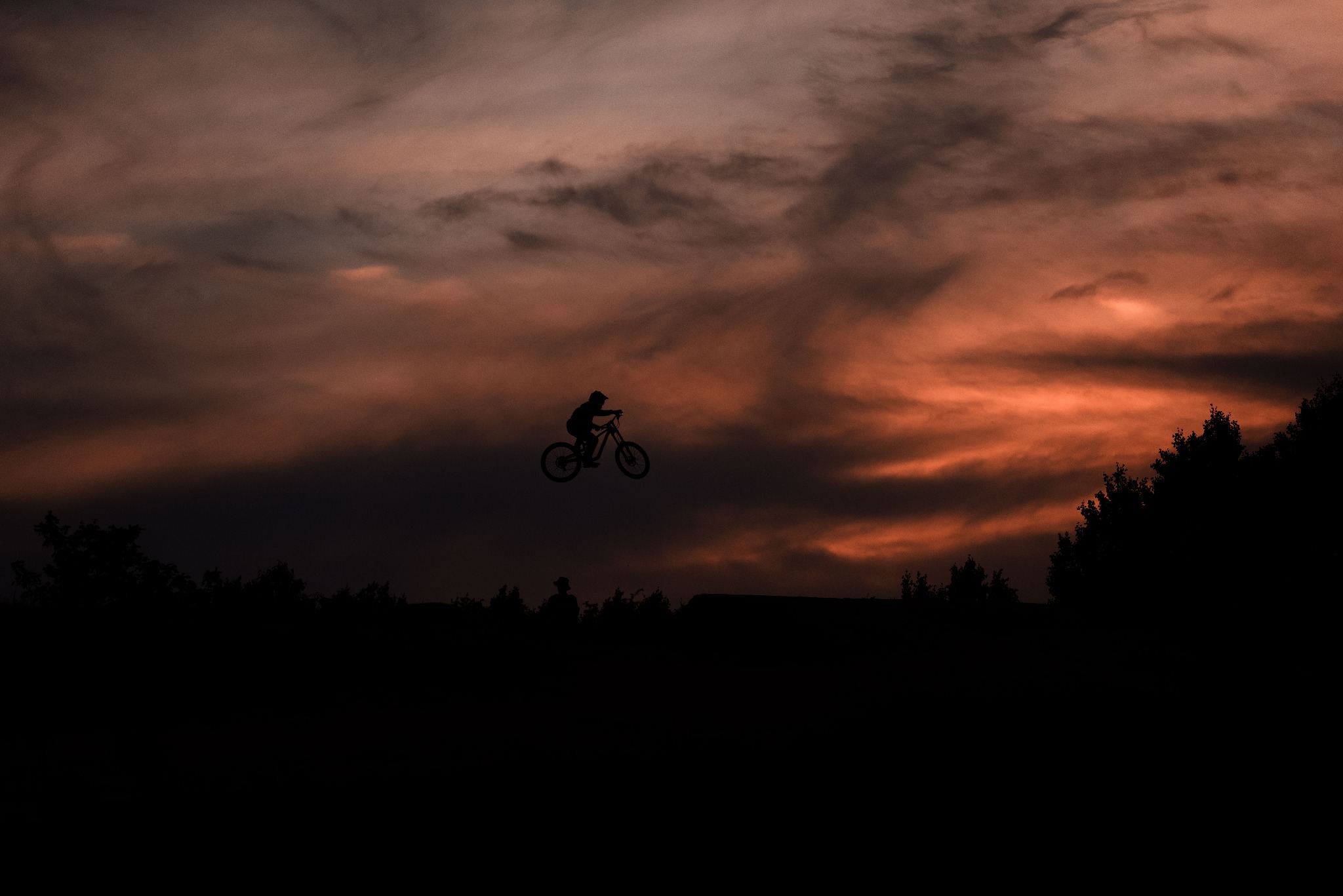 Sylwetka rowerzysty wykonującego skok na tle dramatycznego, pomarańczowo-szarego nieba o zachodzie słońca, zarys lasu w dolnej części kadru.