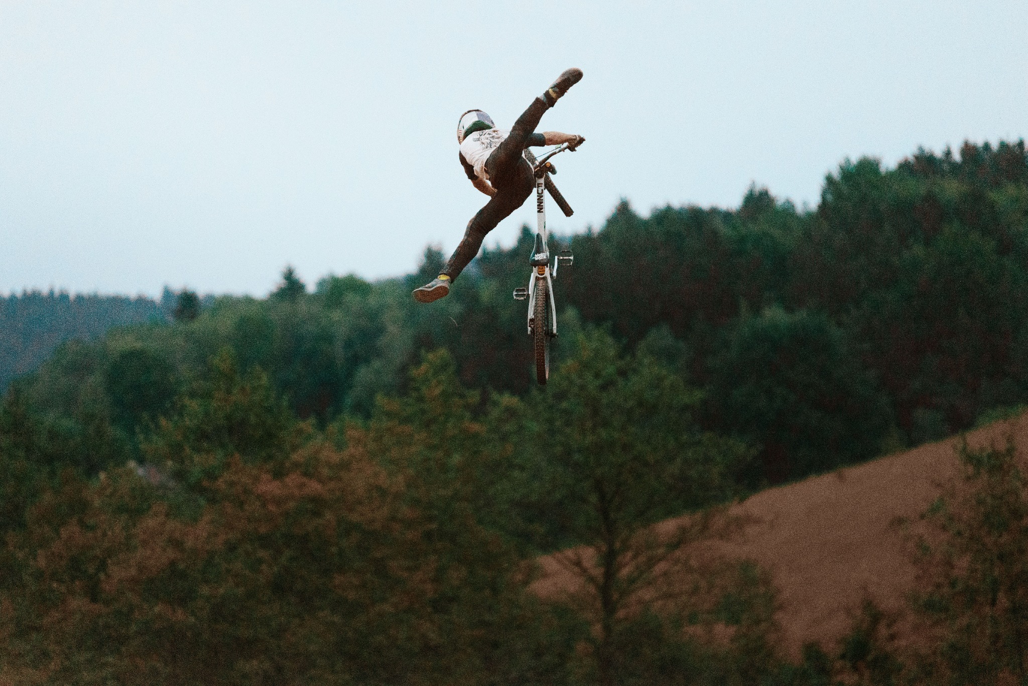 Rowerzysta w kasku wykonuje trik w powietrzu, odrywając się od roweru na tle zalesionego wzgórza.