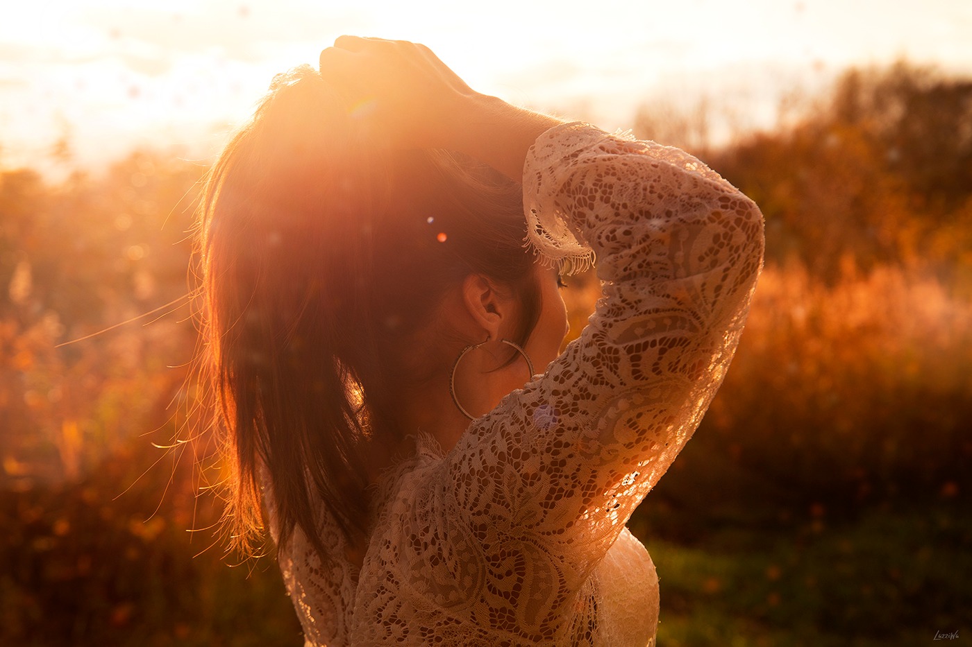 Kobieta w koronkowej bluzce, odwrócona tyłem, poprawia włosy na tle zachodzącego słońca, widoczne kolczyki koła.