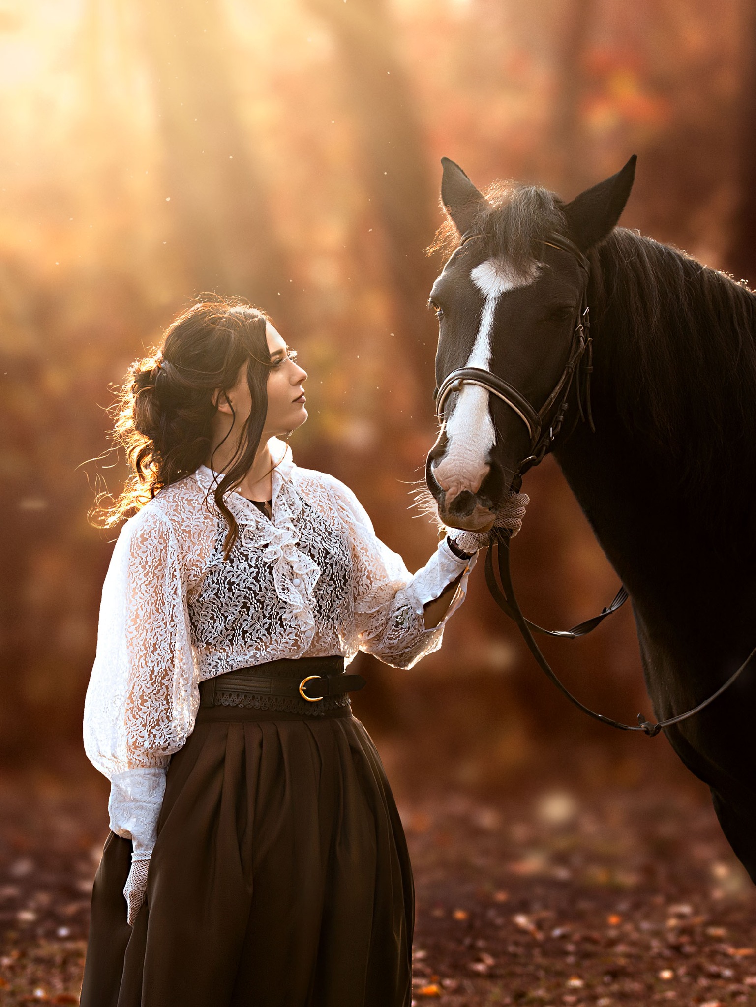 Kobieta w koronkowej bluzce i długiej spódnicy, delikatnie dotyka pyska czarnego konia z białą strzałką na czole, w lesie oświetlonym promieniami zachodzącego słońca.