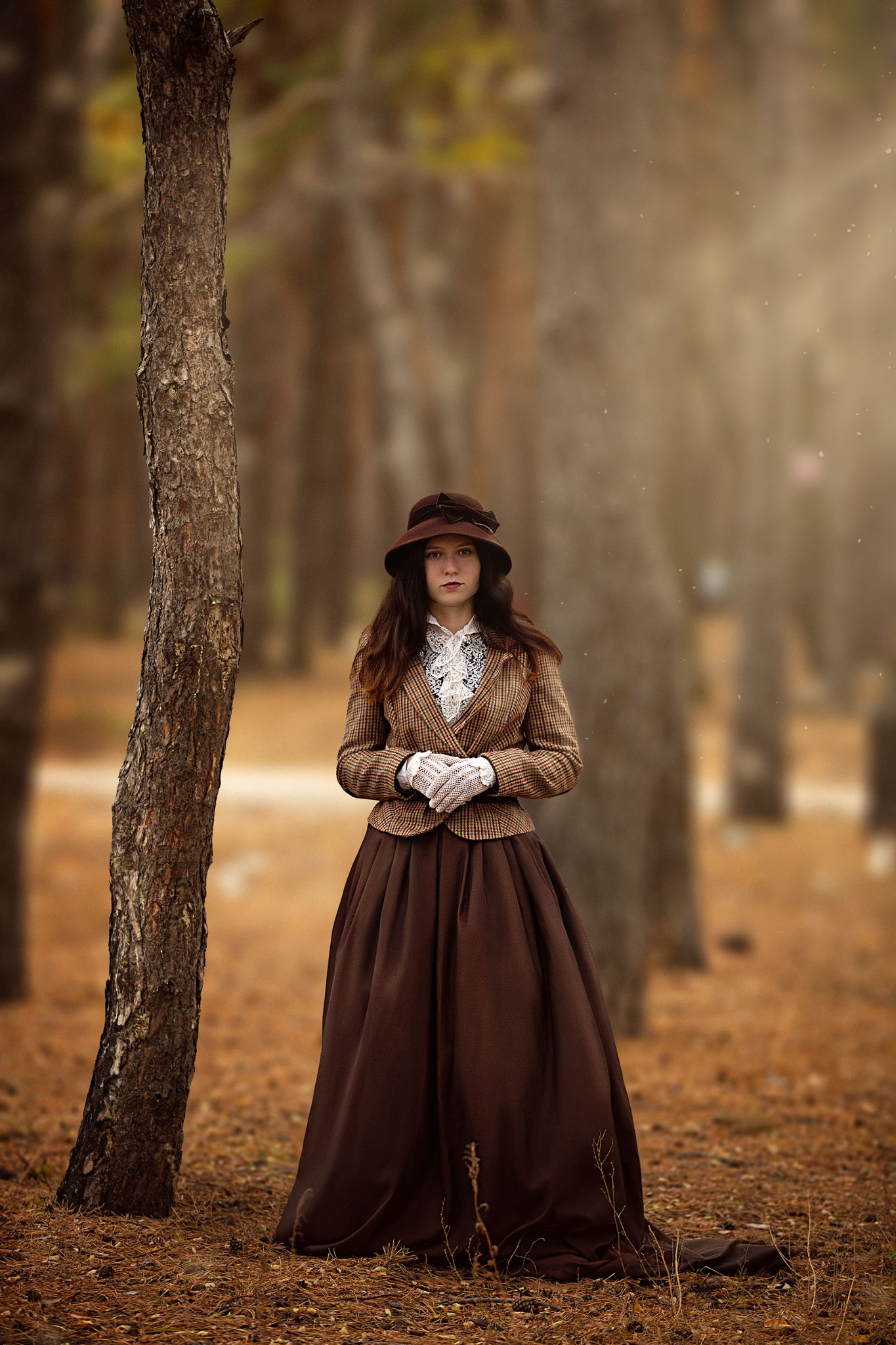 Portret kobiety w brązowej sukni i tweedowej marynarce, pozującej w lesie obok drzewa, z rozproszonym światłem tworzącym subtelną atmosferę.