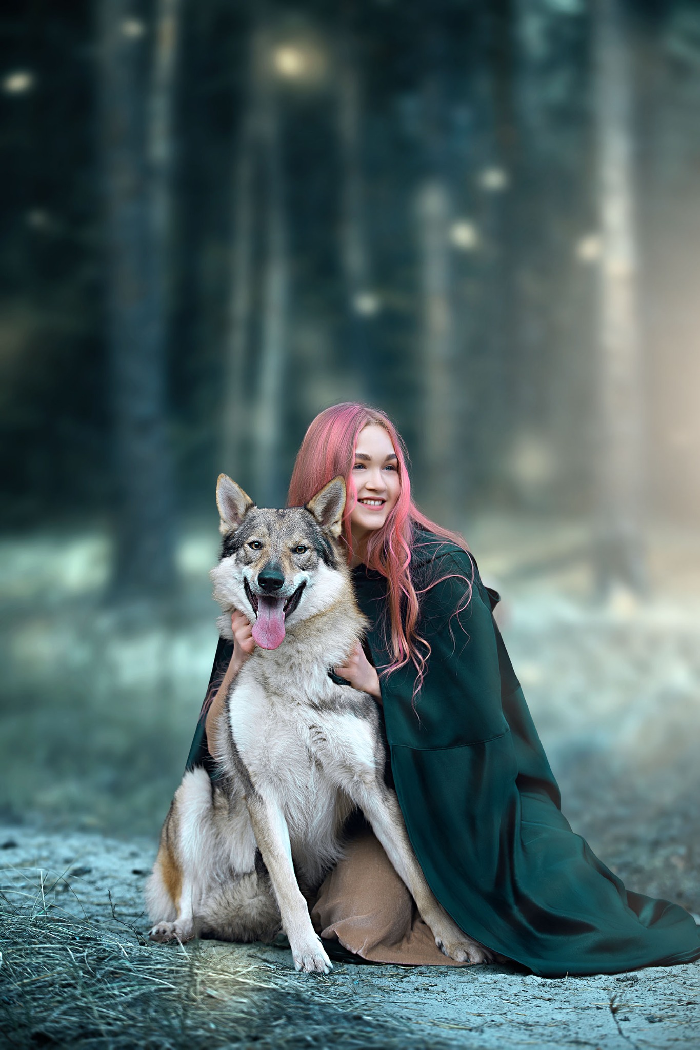 Uśmiechnięta młoda kobieta z różowymi włosami w ciemnozielonej pelerynie obejmuje dużego psa rasy wilczak czechosłowacki siedzącego na tle rozmytego leśnego krajobrazu.