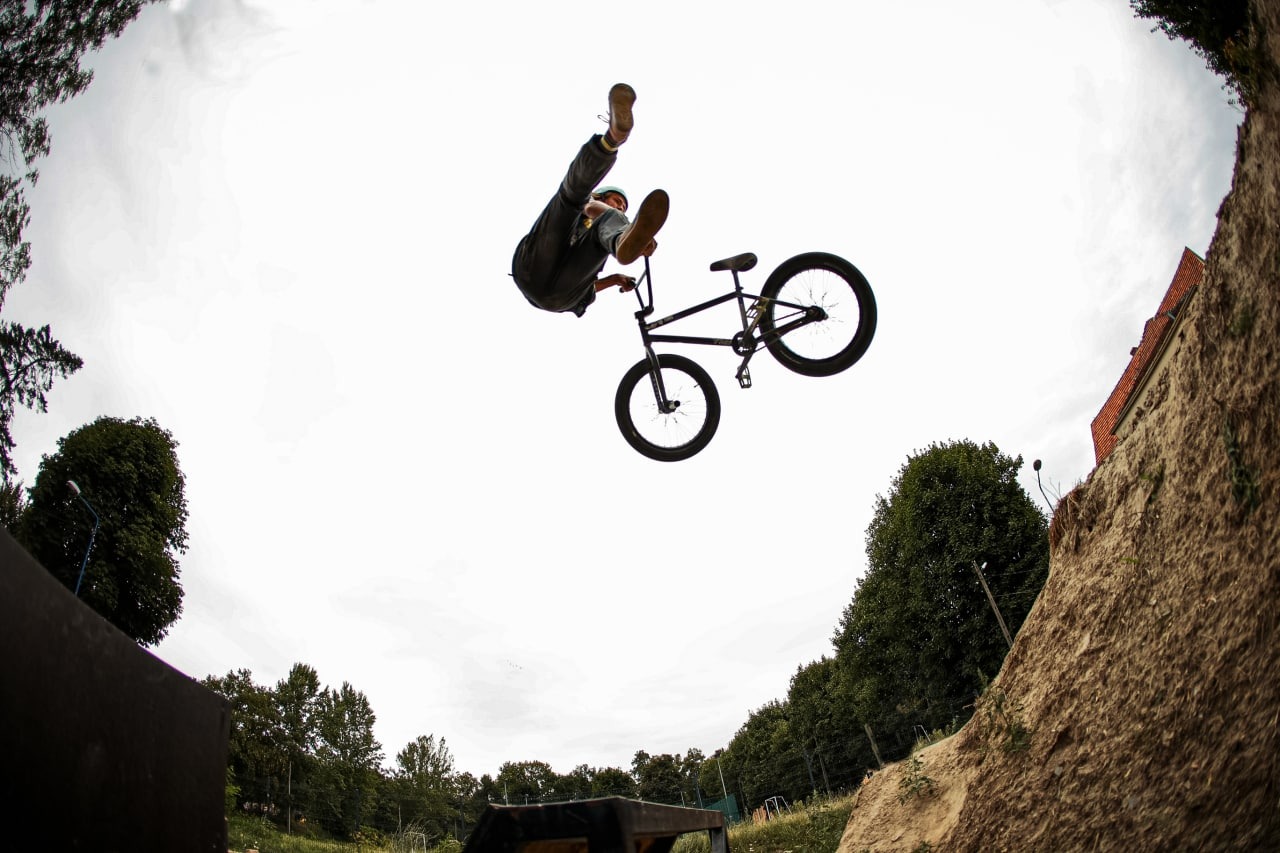 Akrobata na rowerze BMX w powietrzu, widziany z dołu, z ziemnym nasypem i budynkiem w tle.