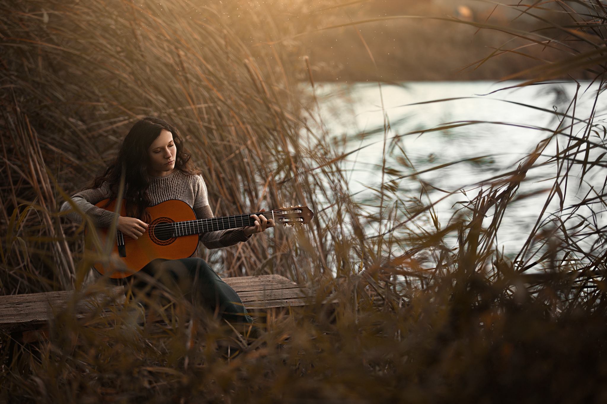 Kobieta z długimi, ciemnymi włosami gra na gitarze klasycznej siedząc na drewnianej ławce pośród wysokich traw w pobliżu zbiornika wodnego w ciepłym, jesiennym oświetleniu.