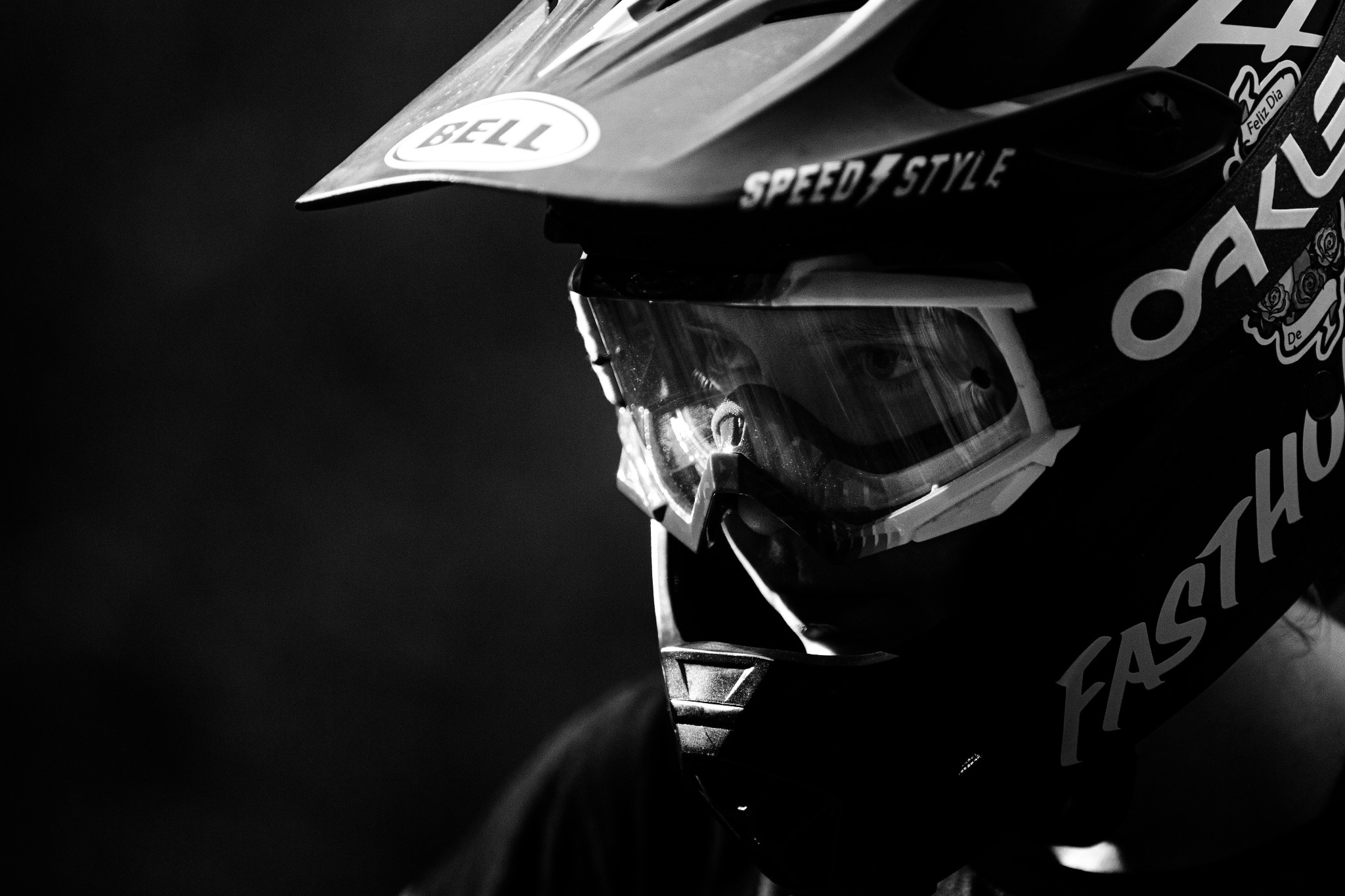 Czarno-białe zbliżenie twarzy osoby w kasku motocyklowym i goglach, widoczne odbicie w goglach, napis SPEED STYLE na kasku, ostre światło boczne.