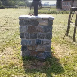 Niska, kamienna kolumna z szarym blatem, wykonana z nieregularnych, szlifowanych kamieni w odcieniach szarości, brązu i zieleni, na tle zielonego trawnika i drewnianego ogrodzenia.