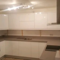 Jasna kuchnia z białymi, lakierowanymi szafkami, szarym blatem i stalowym okapem, oświetlona reflektorami sufitowymi.