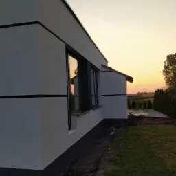 Nowoczesny dom z białą elewacją i czarnymi akcentami w promieniach zachodzącego słońca, widoczne okna z refleksami nieba.